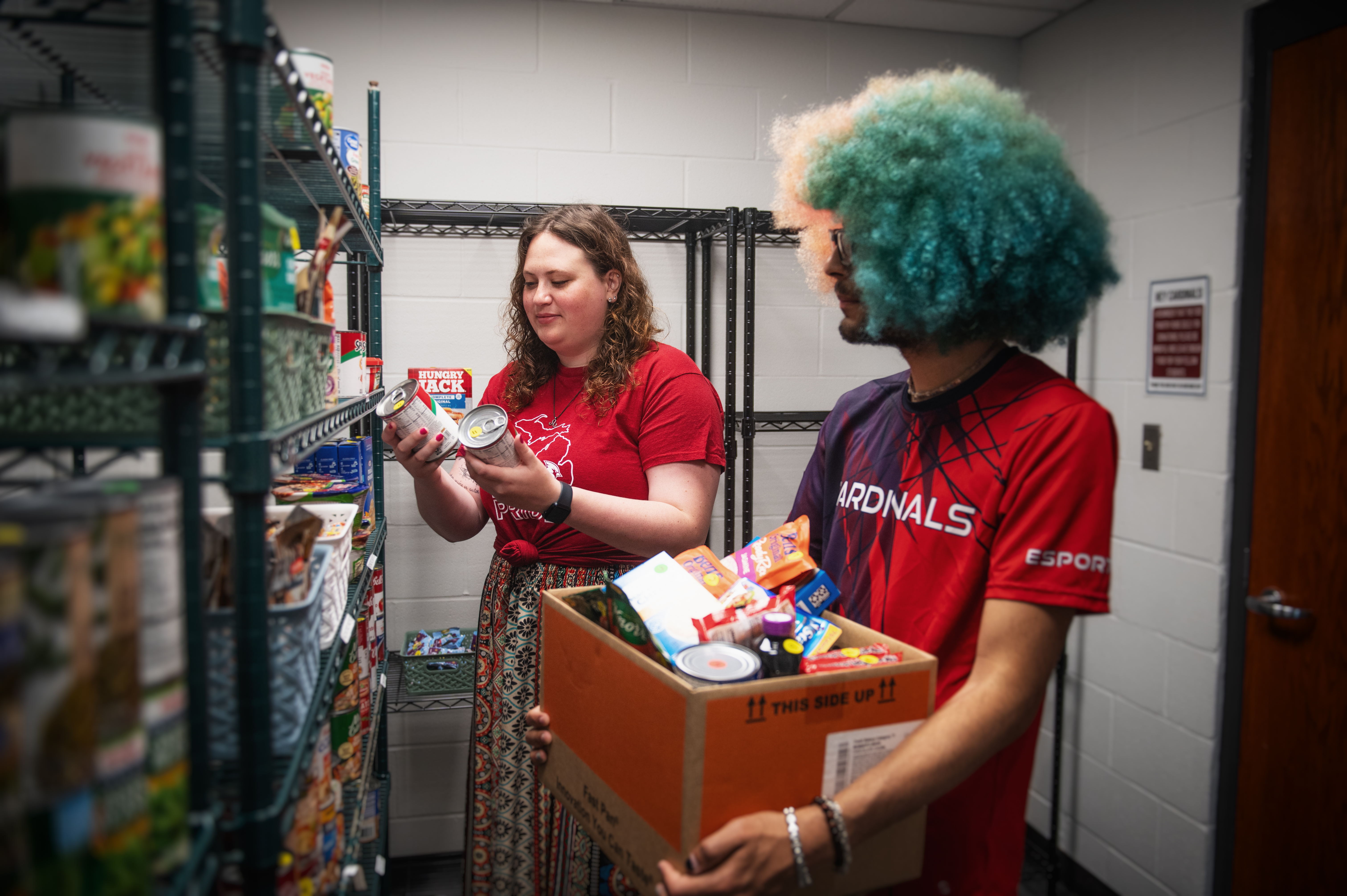 One student holding a box filled with non-perishable food in the Cardinal Food Pantry while the other student add more items to the box.